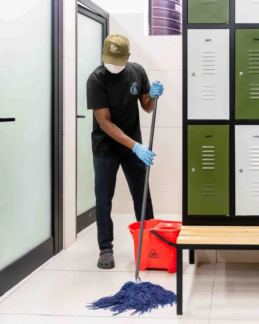 A Clean by AK cleaner mopping a locker room floor beside a wet-floor bucket